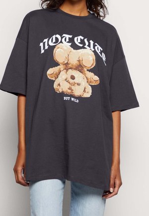 Osoba nosząca czarny oversize T-shirt z beżowym misiem do góry nogami i napisami „NIE SŁODKI” i „ALE DZIKI” z przodu.