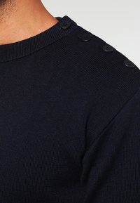 Pull en tricot bleu marine avec un col rond, comportant quatre boutons noirs le long de l'épaule et un tissu texturé.