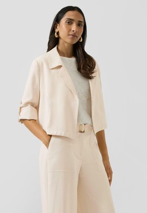 Vrouw met lang donker haar, goudkleurige creolen, een beige cropped jack, een wit kanten topje en beige broek met hoge taille, met handen in de zakken.