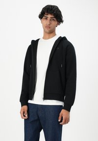Ung mann med krøllete mørkt hår, iført en svart hettejakke med glidelås over en hvit t-skjorte og blå jeans, stående mot en enkel hvit bakgrunn.