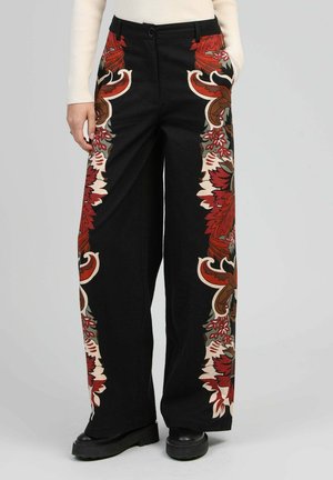 Pantalon large noir avec des motifs floraux audacieux rouges, bruns et beiges le long des coutures extérieures, porté avec des chaussures noires et un pull côtelé crème.