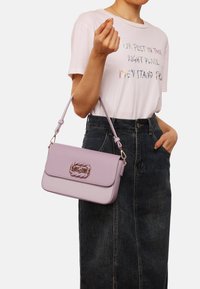 Borsa a tracolla in ecopelle lilla con chiusura a twist-lock, accentata da una treccia e dotata di tracolla rimovibile, abbinata a una maglietta grafica e a una gonna di jeans.