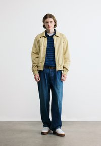 Νεαρό άτομο στέκεται, φορώντας μπεζ μπουφάν, μπλε ριγέ πόλο, άνετο μπλε τζιν και λευκά αθλητικά παπούτσια σε γκρι πάτωμα με απλό φωτεινό φόντο.