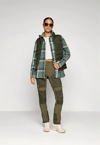 Grønn puffervest over en blå rutete skjorte, kombinert med olivengrønne bukser. Modell iført solbriller. Myke teksturer, funksjonelt design, avslappet utseende.