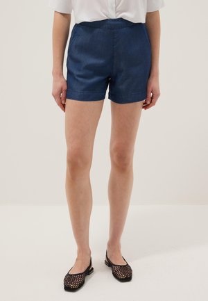 Donna che indossa pantaloncini di jeans blu scuro, top bianco e ballerine nere in rete con cinturino sul retro, in piedi davanti a uno sfondo chiaro e uniforme.