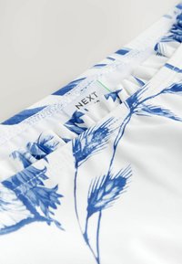 Textil alb cu un model floral albastru, cu margini volăntate. Etichetă vizibilă cu marca "NEXT." Textură netedă cu o ușoară elasticitate.