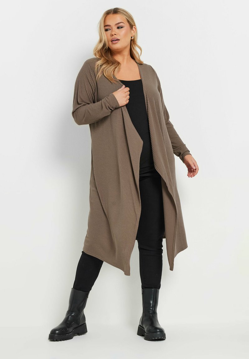 Lång, ribbad kofta i en dämpad olivgrön färg, öppen framtill, med långa ärmar. Kombination med en svart topp och smala svarta byxor, samt kraftiga svarta stövlar.