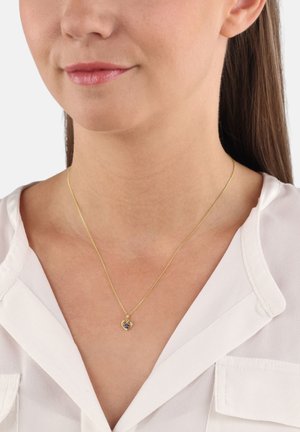 Vrouw die een gouden hartvormige pendantsieraad met een kleine paarse edelsteen draagt, gekleed in een witte V-hals blouse.