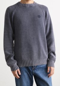 Mann in einem strukturierten dunkelgrauen Strickpullover und blauen Jeans, der mit entspannten Händen an den Seiten vor einem schlichten Hintergrund steht.