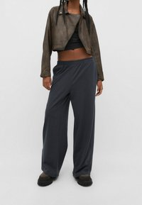 Pantalones de pierna ancha color gris oscuro con cintura elástica, combinados con un top negro corto y una chaqueta de cuero marrón, con cremalleras y un acabado desgastado.