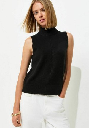 Donna con capelli biondi lisci che indossa un maglione nero a collo alto senza maniche e pantaloni bianchi, con una mano in tasca e un braccialetto d'oro.