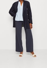 Blazer doppiopetto blu navy, blusa azzurra chiara con motivo a occhio di pernice, pantaloni larghi scuri, ciabatte color crema con dettagli intrecciati.