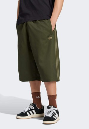 Osoba nosząca oliwkowo zielone długie spodenki Adidas, brązowe skarpetki Adidas oraz czarne buty Adidas z białymi paskami stoi na jednolitym tle.
