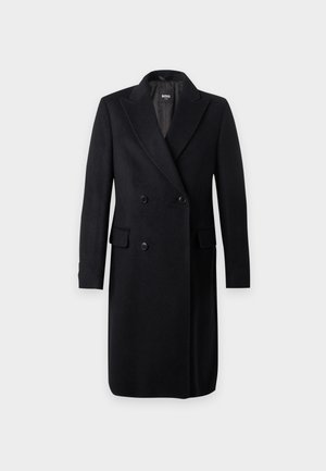 Jeune homme portant un long manteau noir croisé, un pull noir, un pantalon blanc large et des chaussures noires, debout devant un fond blanc uni.