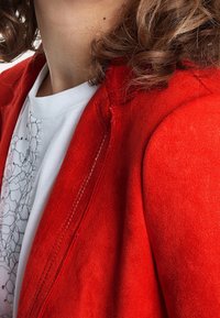 Czerwona zamszowa kurtka z widocznym przeszyciem, dopasowany krój, gładka tekstura i kołnierz z wycięciem, noszona na białej bluzce z ozdobnym wzorem.
