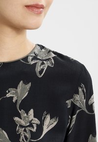 Svart tygtop med grå blommönster, med rundad halsringning och slät textur. Närbild visar sömnadsdetaljer.