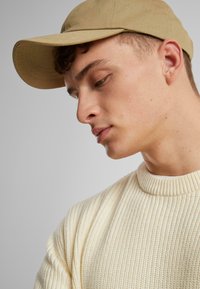 Gorra beige con visera curva, llevada en un ángulo. Suéter de punto acanalado en color crema claro, texturizado con un cuello alto. Tono de piel sutilmente visible.