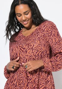 Robe à manches longues dans un motif abstrait bordeaux et orange, en tissu doux avec des poignets froncés et un design en v. Anneaux décoratifs aux mains.