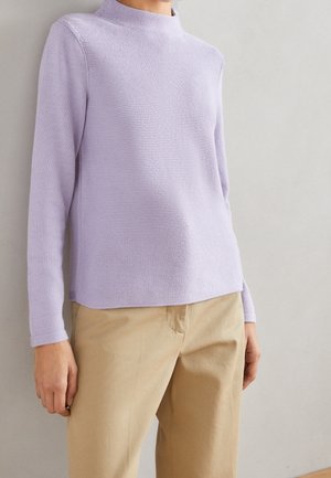 Personne portant un pull texturé lavande à manches longues et un pantalon beige sur un fond gris clair uni.