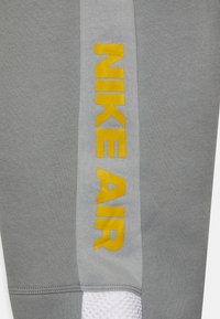 Grå atletiska byxor med en framträdande gul "NIKE AIR"-logotyp, och en strukturerad vit mesh-accent längs fållen för kontrast.