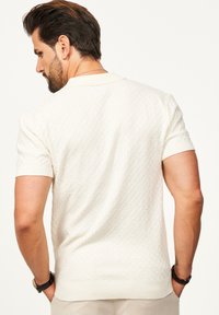 Cremefarbenes Poloshirt mit kurzen Ärmeln aus strukturiertem Material, mit einem gerippten Kragen und Abschlüssen. Verfügt über ein geometrisches Muster für zusätzliche Details.