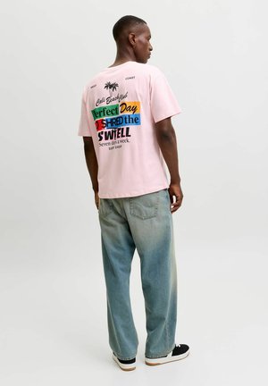 Homme portant un t-shirt graphique rose et un jean bleu ample, debout de dos devant un fond uni.