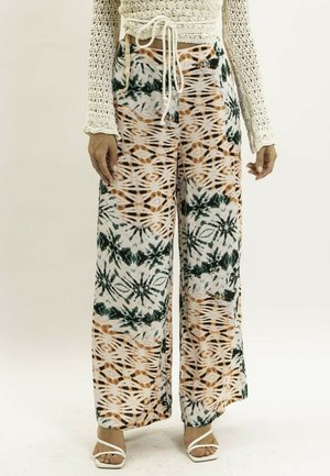 Femme portant un pantalon à jambes larges avec un motif tie-dye orange et vert, un pull en crochet crème et des sandales blanches à brides.