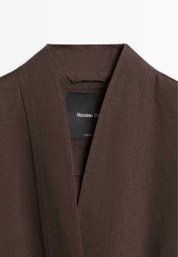 Close-up van de halslijn van een bruin kledingstuk met een V-vormige kraag en een zwart label aan de binnenkant met de merknaam.