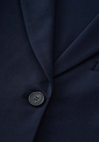 Navy blue blazer featuring a notched lapel, smooth fabric texture, and a black button with four holes near the lapel for closure.