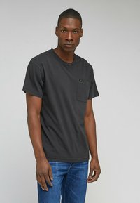 Svart bomullströja med rund hals, korta ärmar och en ficka i fram. Har en diskret logotagg på fickan. Bärs med blå jeans.