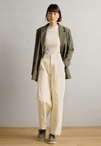 Blazer verde oliva, maglietta bianca a coste, pantaloni larghi color avorio e scarpe grigie. Texture lisce e design minimalista.