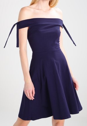 Frau trägt ein marineblaues schulterfreies Kleid mit gebundenen Trägern an den Armen und einem ausgestellten knielangen Rock.