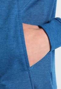 Blå, texturerad tyg med en ficka vid midjan. Närbild av en hand som vilar inuti fickan, som visar materialets mjukhet.