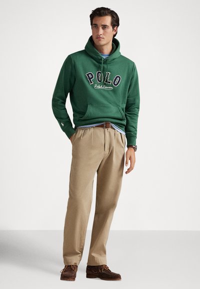 Vyras dėvi žalią Polo su gobtuvu, smėlio spalvos klostuotas kelnes su rudu diržu, rudi odiniai batai, stovi priešais baltą foną.