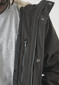 Mann, der eine teilweise offenstehende schwarze Jacke mit Fleece-Innenfutter über einem hellgrauen Rundhalshemd trägt.