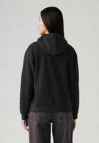 Sudadera negra con un bolsillo tipo canguro en el frente, capucha con cordón y puños acanalados. Combinada con pantalones de mezclilla grises que presentan un ajuste relajado.