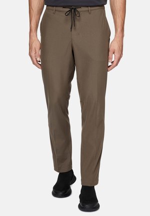 Pantaloni marroni da uomo in tessuto leggero con vita regolabile, gambe a sigaretta e due tasche laterali. Indossati con scarpe nere.
