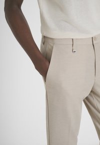 Pantaloni beige su misura realizzati in tessuto testurizzato con un motivo sottile; presentano tasche inclinate e un piccolo dettaglio in metallo argentato.