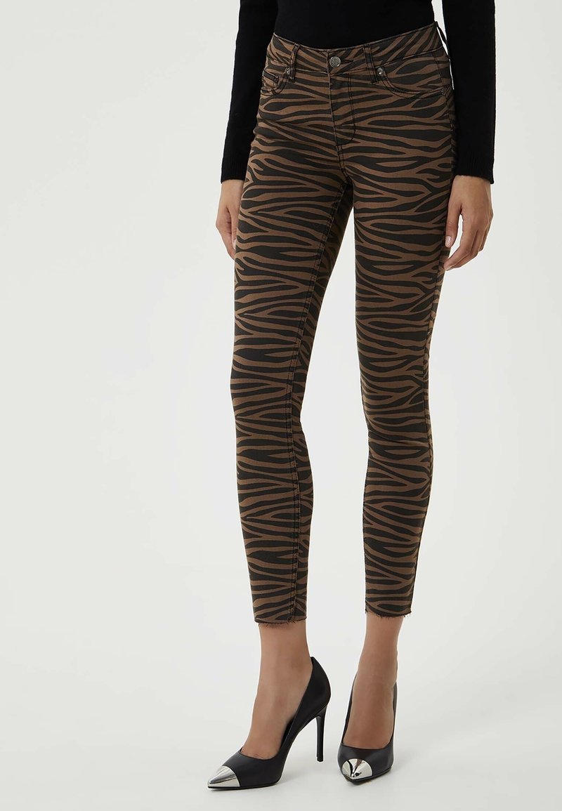 Jeans ajustados de estampado de cebra marrón con acentos negros, que presentan una cintura alta, bolsillos frontales y un dobladillo deshilachado, combinados con tacones negros de punta.