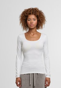 Bílá halenka s dlouhými rukávy a kulatým výstřihem, přiléhavého střihu, hladké textury, bez viditelných vzorů či akcentů. V kombinaci se šedými kalhotami.