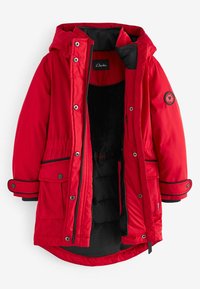 Červená parka bunda s čiernou vnútornou podšívkou, vybavená zipsom na prednej strane, zapínaním na gombíky, nastaviteľnou kapucňou a okrúhlym logom na rukáve.