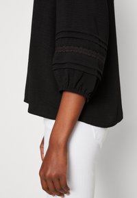 Blouse noire en tissu texturé ; manches trois-quarts avec poignets froncés et accents en dentelle décorative près du coude.