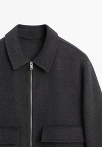 Giacca grigia in misto lana con colletto piatto, chiusura con zip e due tasche frontali. Presenta una finitura testurizzata e una forma strutturata.