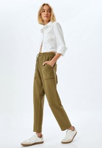 Witte cropped shirt met opgerolde mouwen, gecombineerd met olijfgroene joggers met grote zijzakken en lichtgekleurde sneakers.