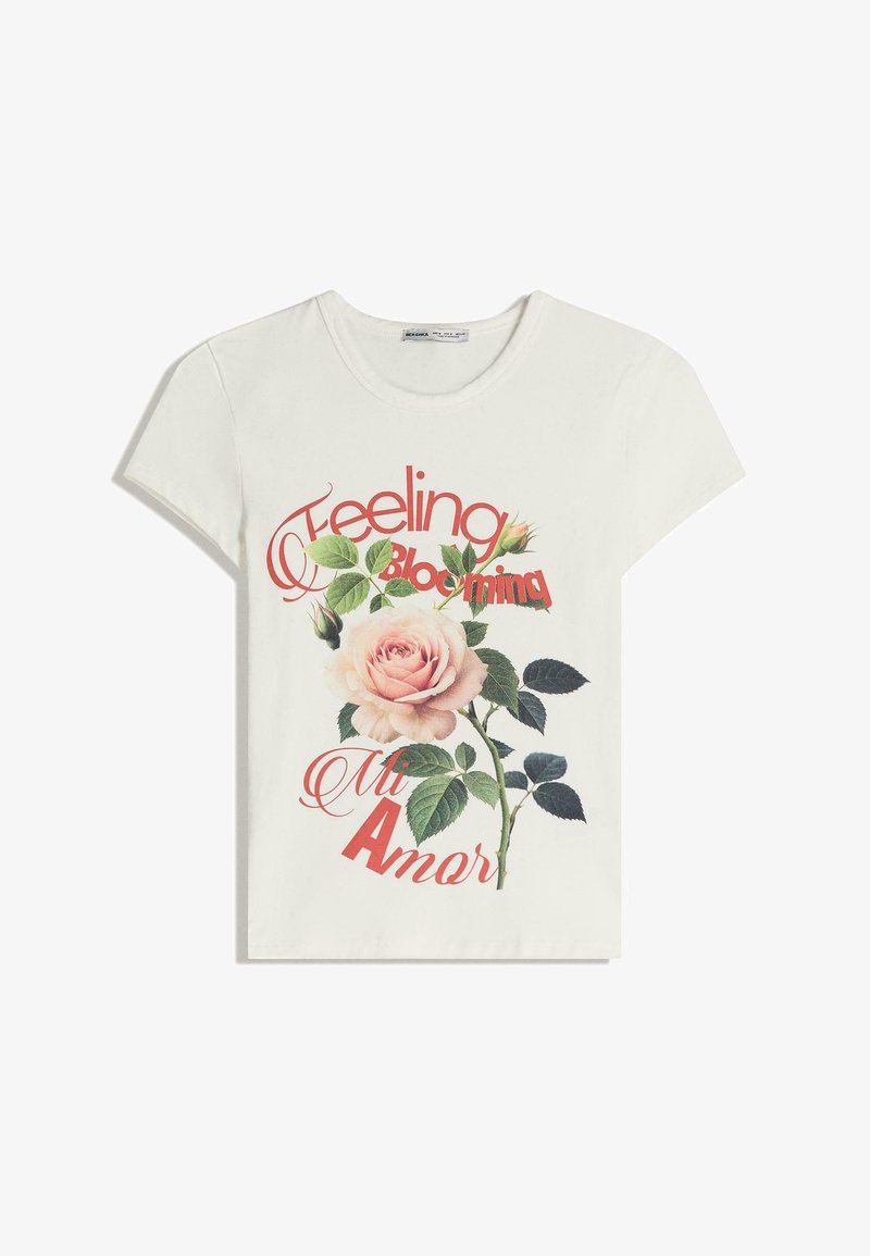 T-shirt bianco in cotone con scollo rotondo. Presenta una grande grafica di una rosa rosa, foglie verdi e testo rosso che dice: "Feeling Blooming Mi Amor."