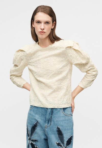 Κρεμ μπλούζα με υφή λουλουδένιου μοτίβου, φουσκωτά μανίκια και στρογγυλή λαιμόκοψη. Συνδυασμένη με μπλε τζιν παντελόνι με κεντημένο σχέδιο φύλλων.