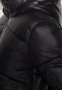 Doudoune noire avec une finition brillante, un motif matelassé et une capuche. Elle possède une fermeture éclair avant visible et une texture douce et rembourrée.