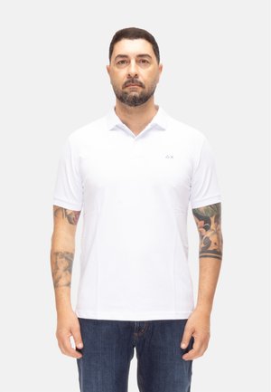 Hombre con el pelo corto y oscuro y barba, viste una camiseta polo blanca lisa y vaqueros oscuros, mostrando tatuajes en ambos antebrazos, de pie frente a un fondo claro.