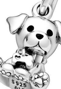 Charm de perro de plata con características faciales y patas detalladas, sosteniendo un hueso con un corte en forma de corazón; acabado pulido y textura brillante.