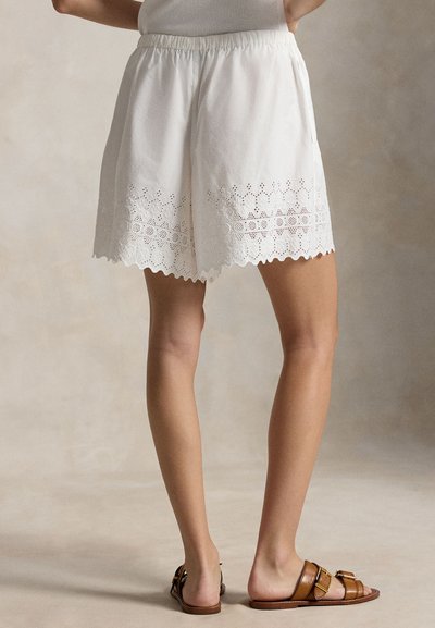 Γυναίκα που φοράει λευκά σορτς με κεντήματα eyelet και κυματιστό τελείωμα, καθώς και καφέ σανδάλια με αγκράφα, στέκεται μπροστά σε ουδέτερο φόντο.
